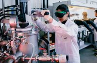 Two researchers in white lab coats operate high-temperature alloy testing equipment inside a materials lab.